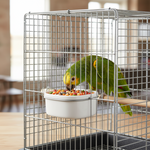 Mixed bird seed in a white bowl inside metal cage, with a green parrot