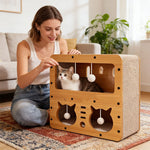 Cardboard cat house with window openings and hanging plush balls; white tabby cat inside