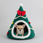 Green plush Christmas tree pet cave bed with white trim, red bow, and pom accents; small dog inside