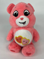 Pink plush dog toy bear with heart-and-sunflower belly graphic and smiling face.