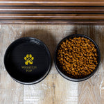 Two black ceramic bowls with gold paw logo; one filled with dry kibble